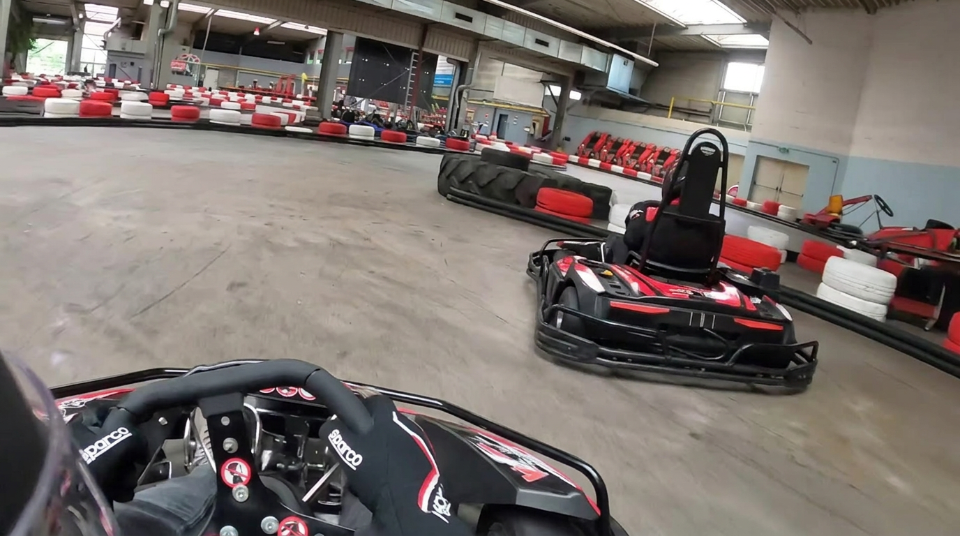 POV-Aufnahme aus einem Kart auf einer Indoor-Kartbahn, im Vordergrund das Lenkrad, davor ein weiteres schwarz-rotes Kart